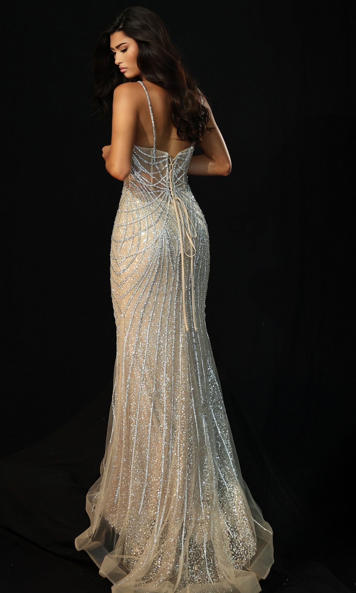A woman poses against a black background wearing the Jovani 49718 long prom dress, featuring sparkling silver fabric, thin straps, a corset bodice, and lace-up back detailing.